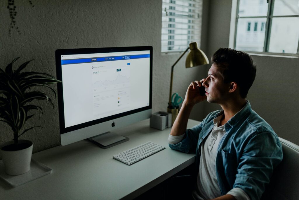 A man in a social media manager role reviews a Facebook page on a monitor while talking on the phone.