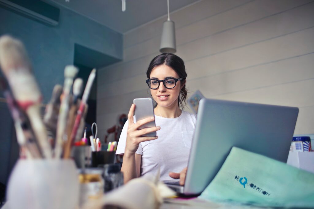A young woman in a social media manager role consults her smartphone while seated at a laptop, with brushes and other creative tools visible in the foreground.