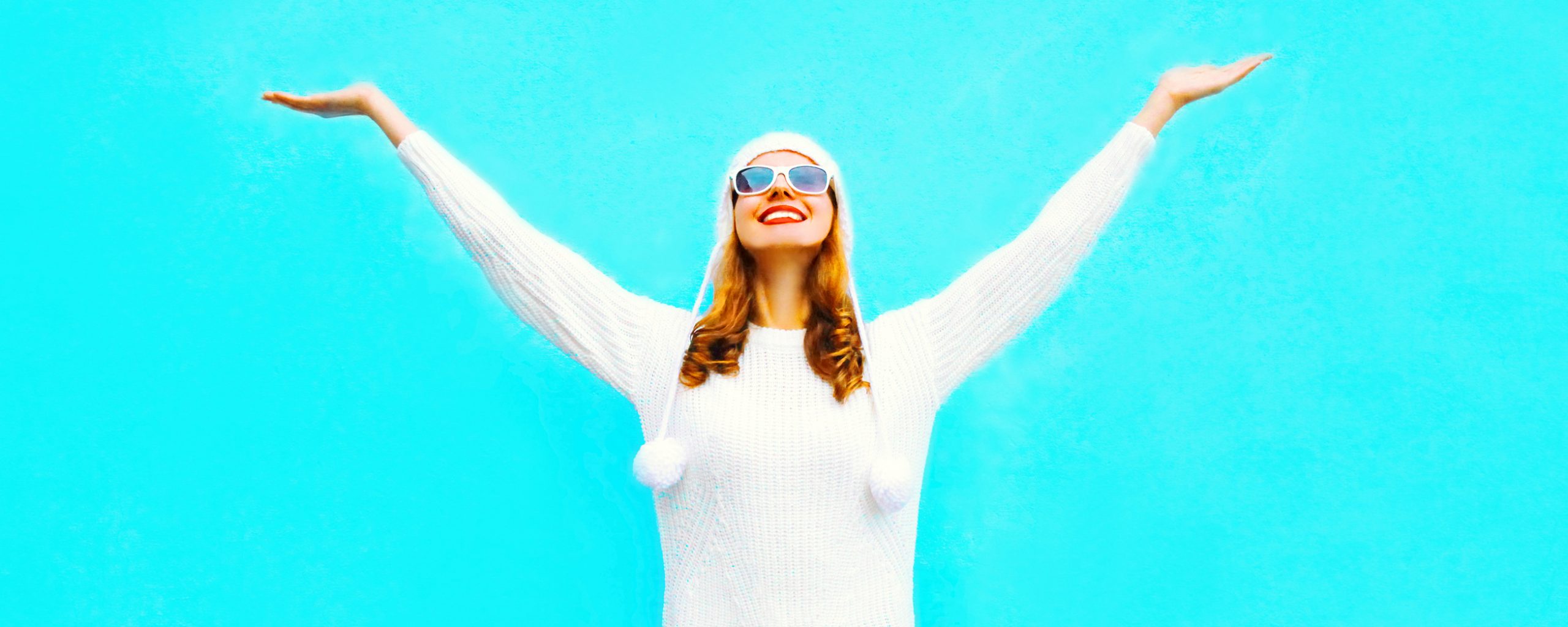 Smiling woman in a white sweater holding her arms up in the air against a blue background.