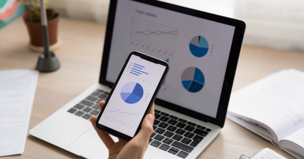 A closeup of a hand holding a smartphone in front of a laptop, both screens show SEO data graphics. 