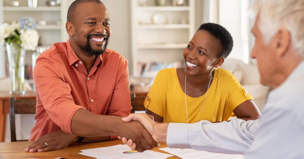 A married couple shakes hands with their wealth management professional across a kitchen table.