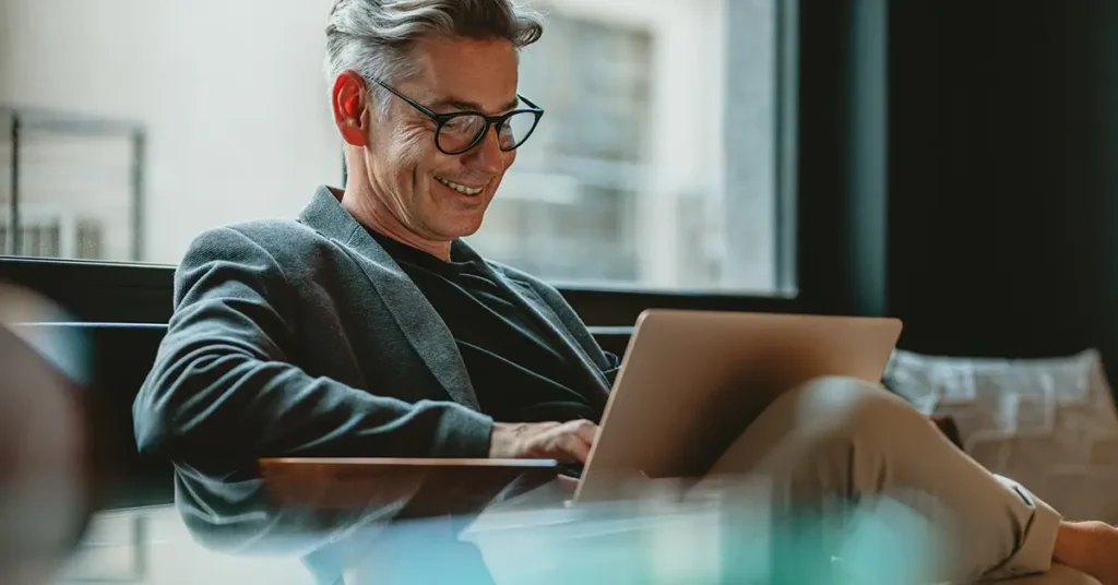 Smiling businessman sitting in office lobby reading great email subject lines on a laptop.